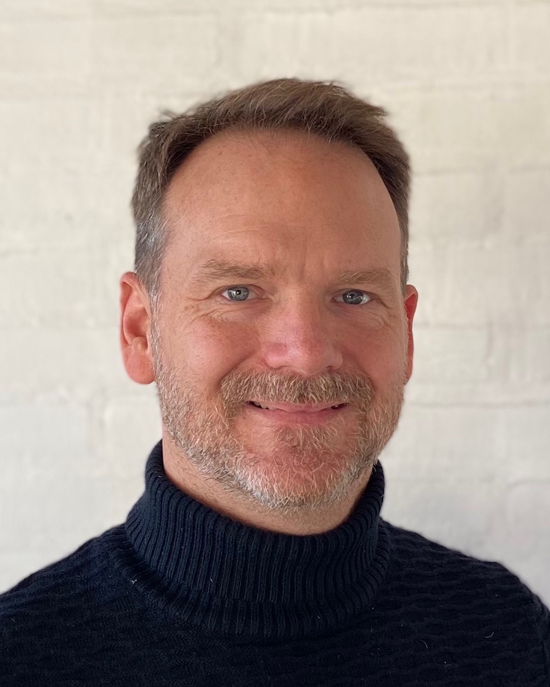 Portrætfoto af underviser på Anger Management kursus og psykoterapeut i Nordsjælland Rasmus Ranmar hos iLivet, i blå rullekrave sweater foran hvid murstensvæg, der tilbyder gestaltterapi gennem gestaltteater og psykoterapi i Nordsjælland og angstbehandling, hjælp til stress, livscoaching og coaching i Nordsjælland